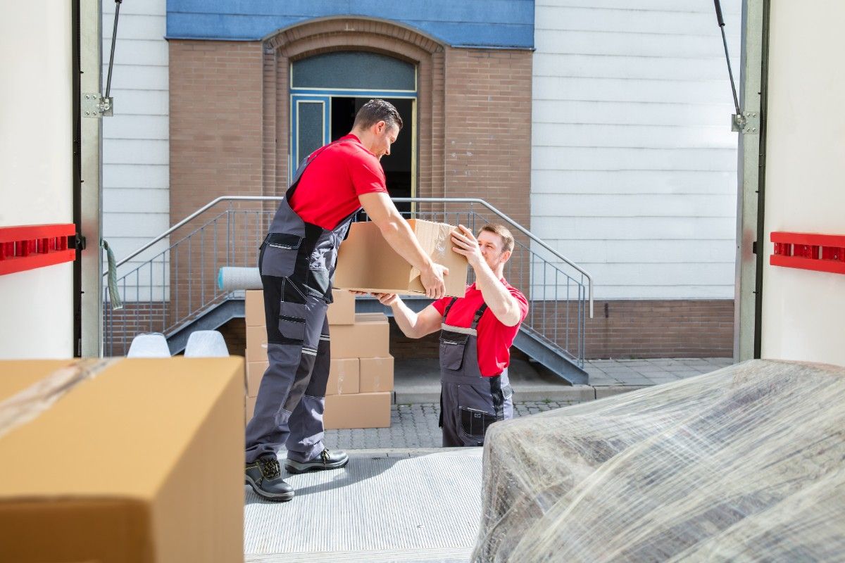 Two Denver movers and packers in red shirts are unloading boxes from a moving truck onto the ground.